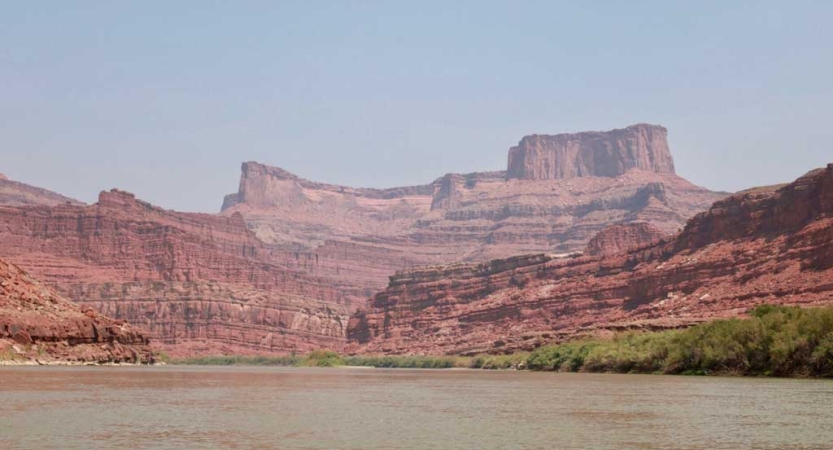 A calm river flows in front of a vast and tall red rock formation. 